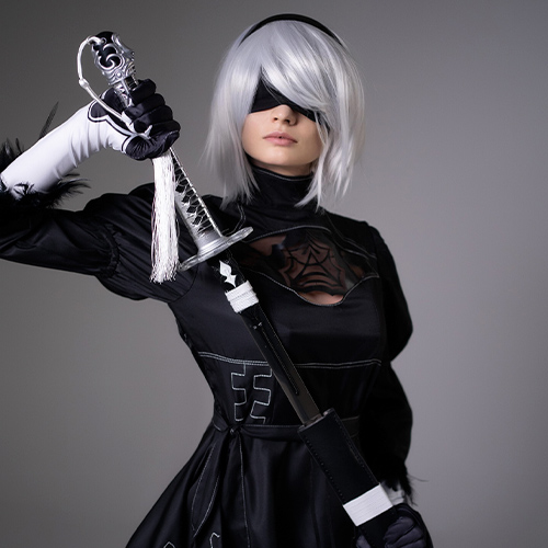 Cosplayer dressed as 2B from Nier: Automata stands confidently, holding a katana over her shoulder. She is wearing 2B's iconic black dress with intricate white embroidery, a blindfold covering her eyes, and short white hair, perfectly capturing the character's mysterious and powerful presence.