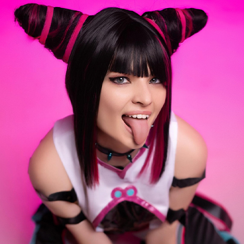 Playful Juri Han cosplay from Street Fighter, crouching with her tongue out, showcasing her bold black and pink hair and gloves in a vibrant pink-lit studio