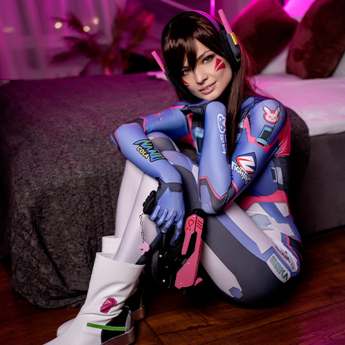 A stunning D.Va cosplay from Overwatch, showcasing the character's vibrant bodysuit and iconic bunny-emblem accessories. The gamer-inspired pink and blue suit highlights every detail, from the headset to the futuristic blaster. Captured in a relaxed pose on a hardwood floor with a soft pink-lit background, this portrayal emphasizes D.Va's playful yet focused personality. Perfectly executed for fans of Overwatch and gaming-themed cosplay.