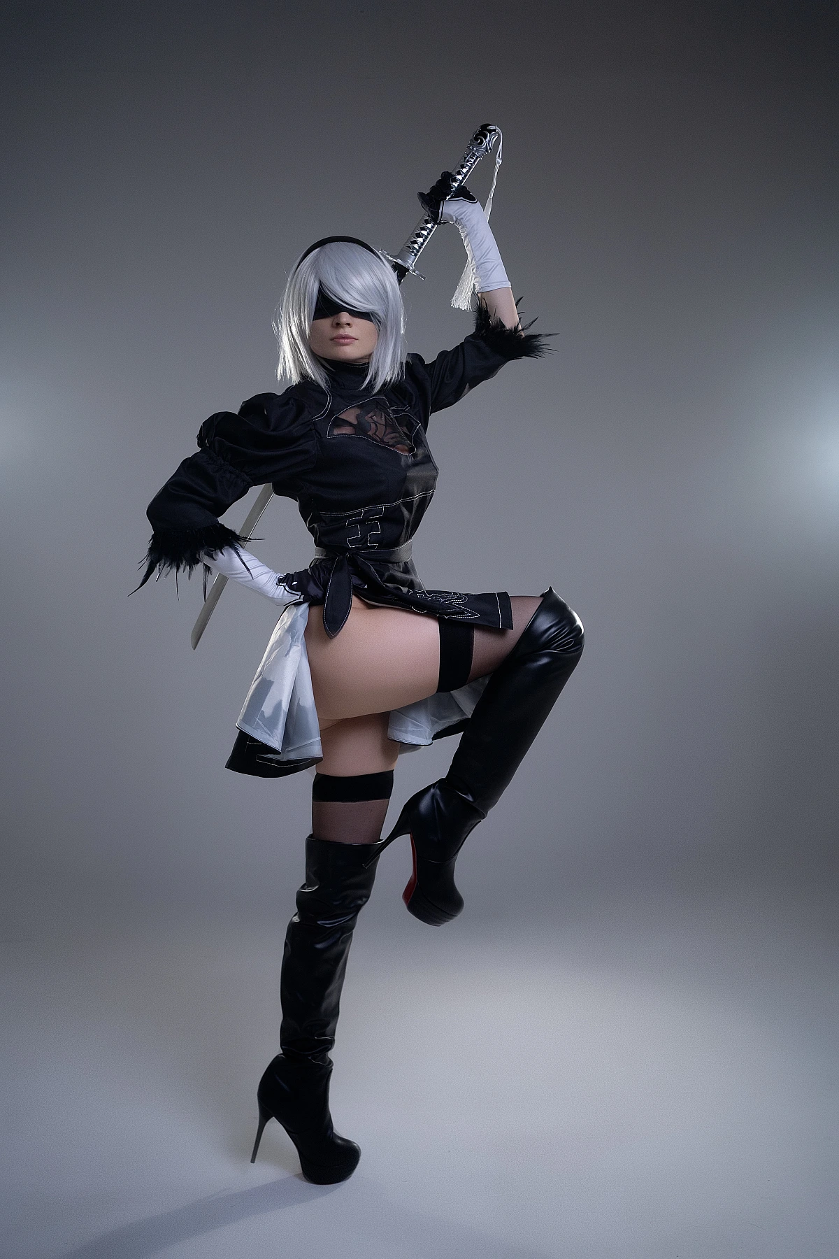2B NieR Automata cosplay dynamic pose with raised leg and sword, silver hair, blindfold, black YoRHa dress and boots