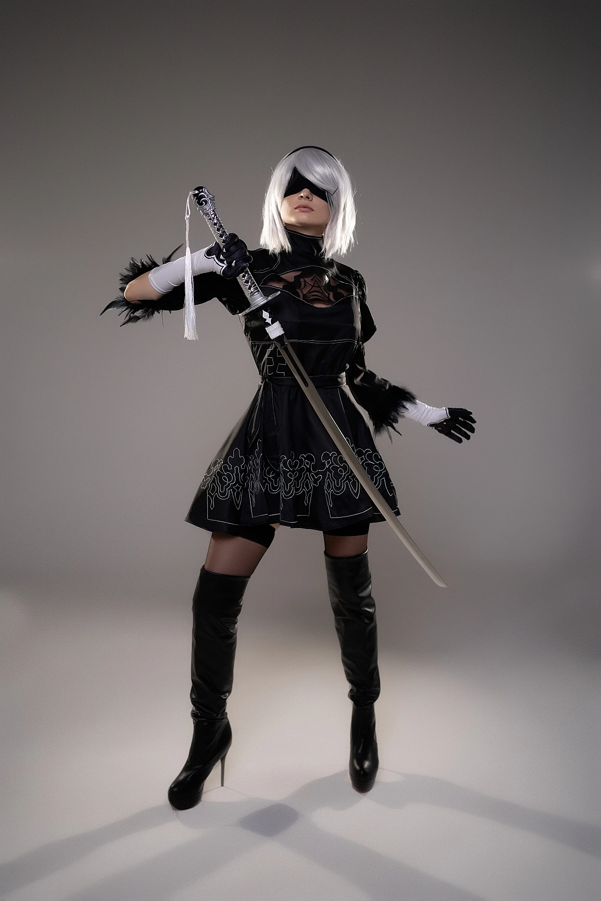 2B YoRHa cosplay frontal pose with katana extended forward, silver hair, black blindfold, NieR Automata costume in studio