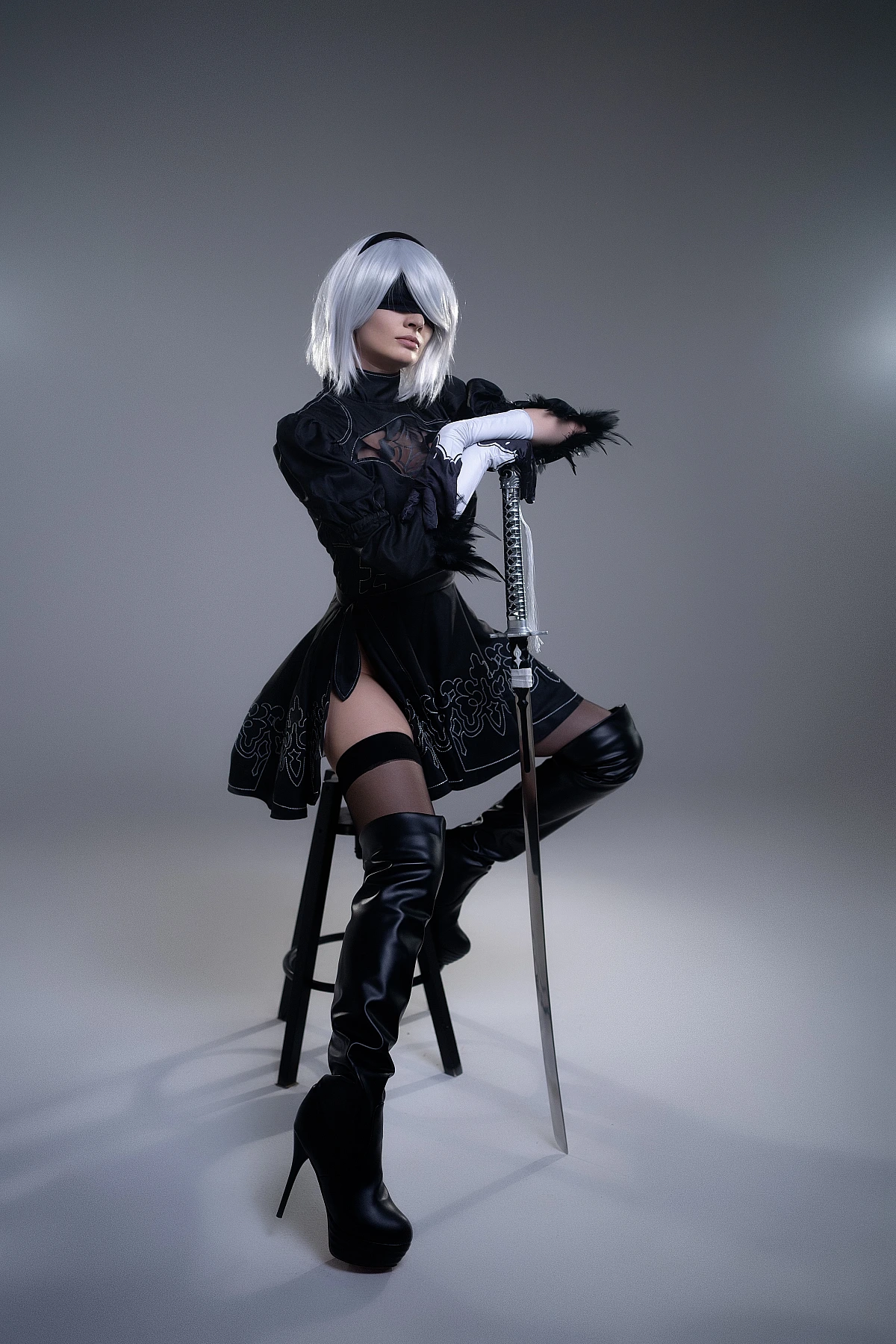 2B NieR Automata cosplay seated portrait with katana, silver hair, black blindfold, YoRHa black dress and thigh-high boots in studio