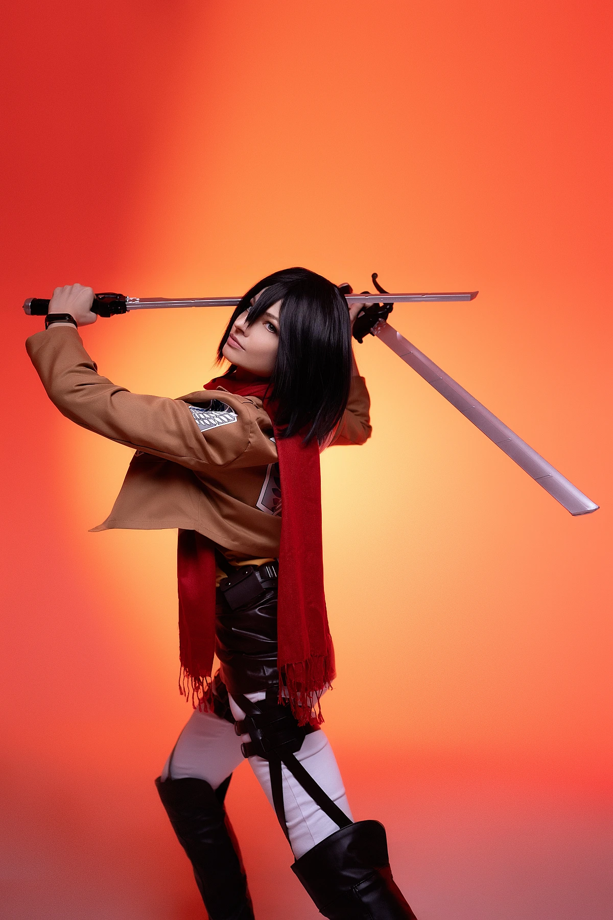mikasa ackerman action cosplay pose with dual odm blades behind shoulders and red scarf against orange backdrop