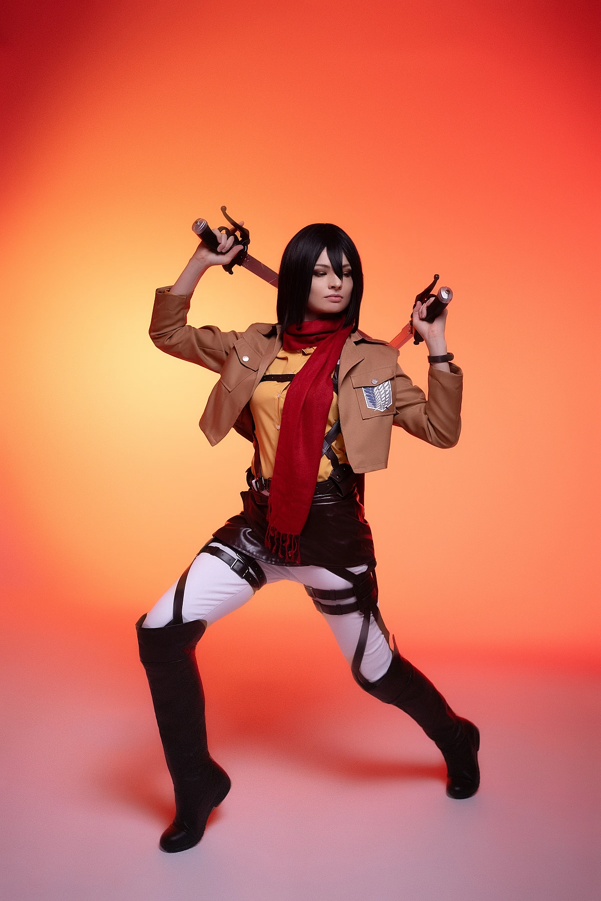 mikasa ackerman cosplay low combat stance with dual blades near shoulders and red scarf against orange studio lighting