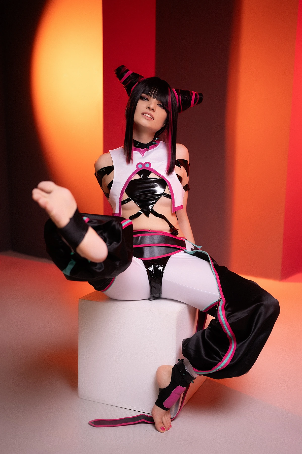 Juri Han Street Fighter cosplay seated on white cube with barefoot leg raised, black strap costume, red-orange neon studio backdrop