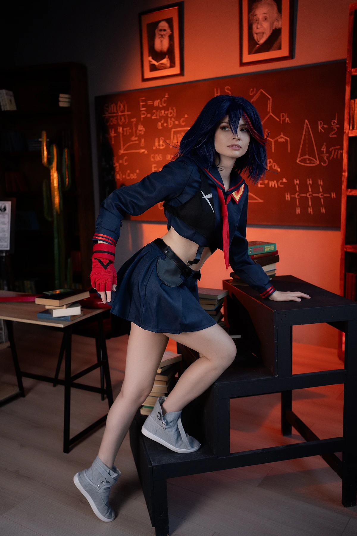 Ryuko Matoi cosplay sitting on stool by desk with textbooks in warm orange light