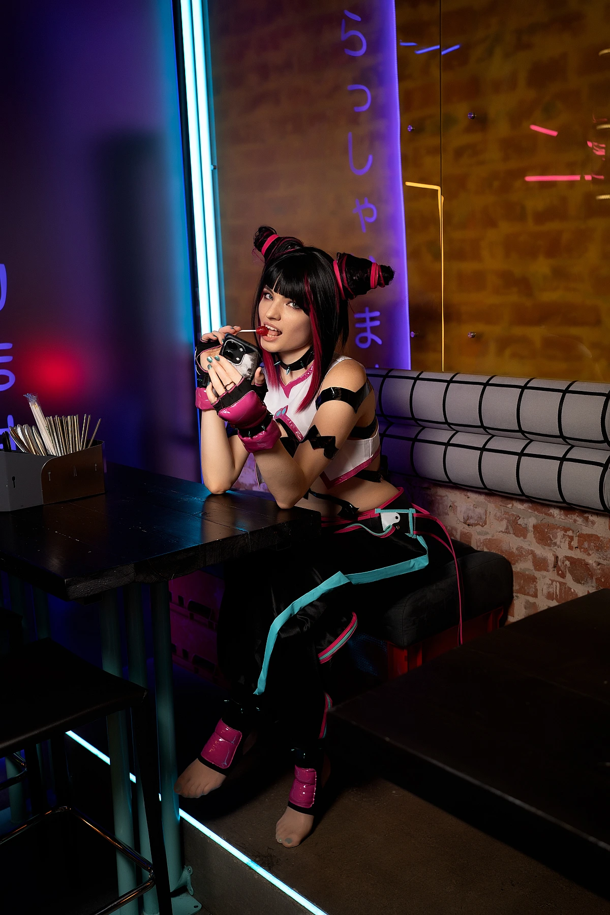 Juri Han cosplay seated at bar table, leaning forward with confident pose, violet-blue neon lighting and barefoot styling
