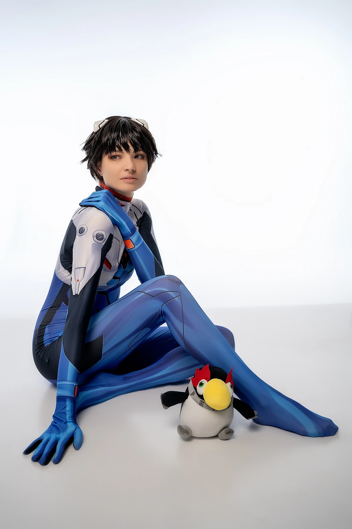 Shinji Ikari cosplay floor scene with plush toy, blue catsuit, extended legs, white studio backdrop, anime cosplay