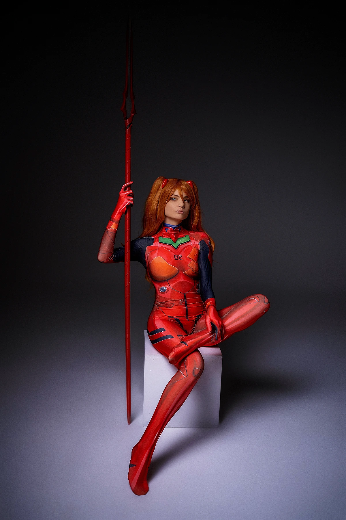 Asuka Langley cosplay seated with crossed legs holding vertical weapon in red plugsuit