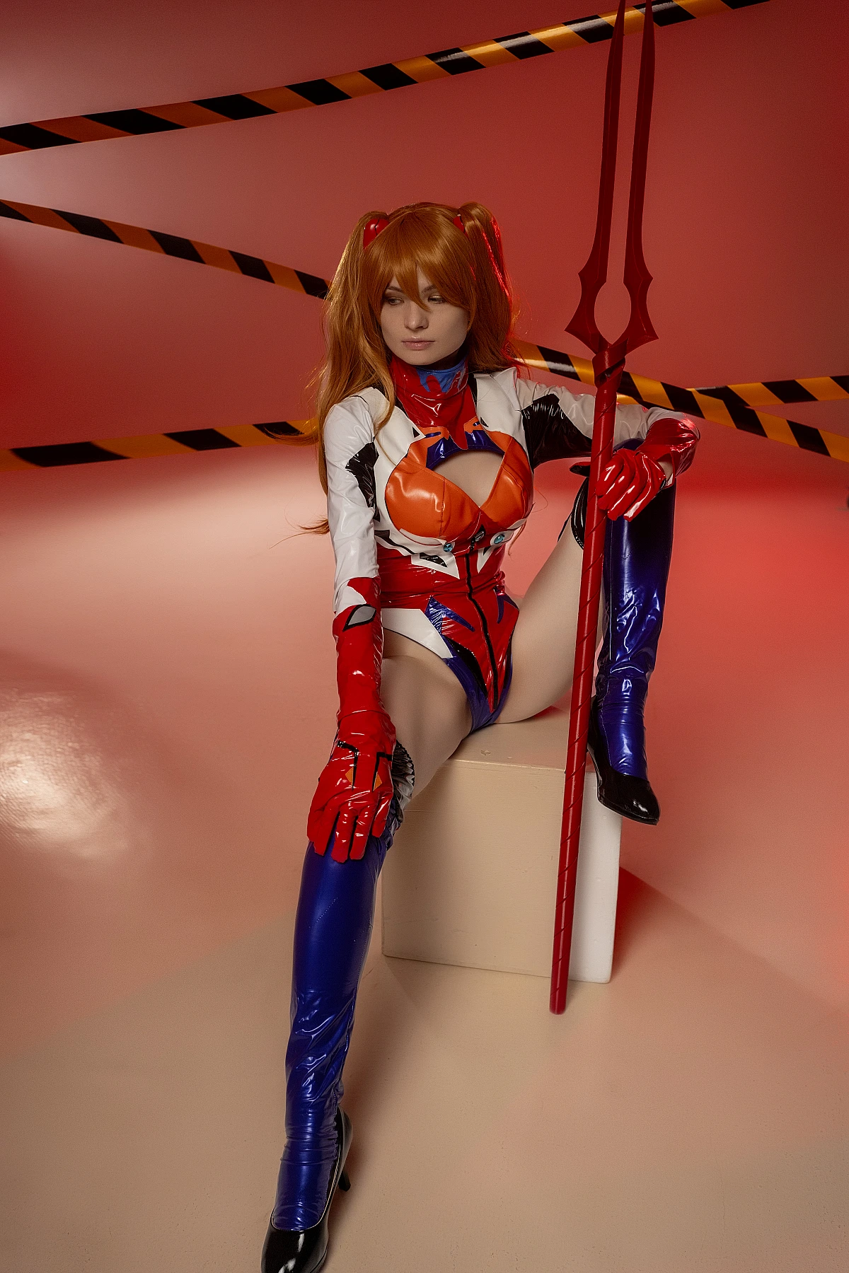 Androgynous Asuka femboy cosplay seated with spear in latex plugsuit red background