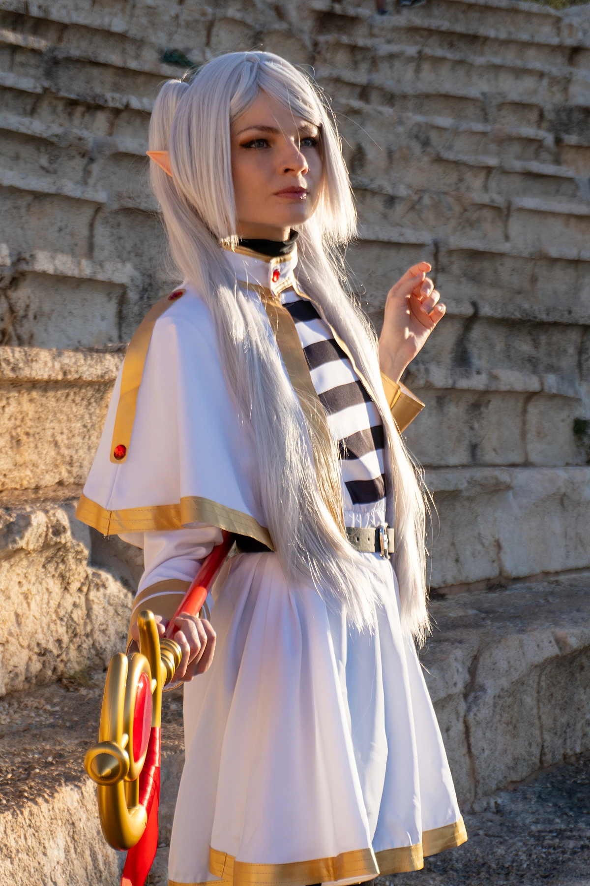 Frieren cosplay half-turned pose staff in hand silver hair elf ears white gold mage dress weathered stone background