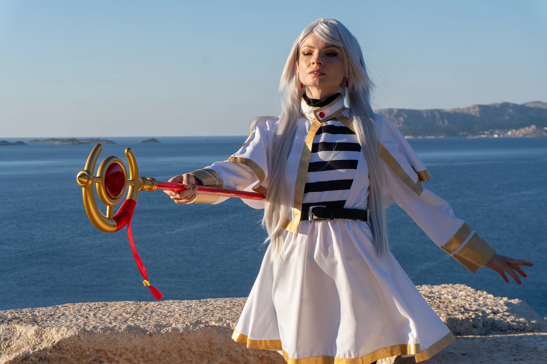 Frieren cosplay frontal stance facing sea staff extended outward silver hair elf ears white gold dress coastal landscape