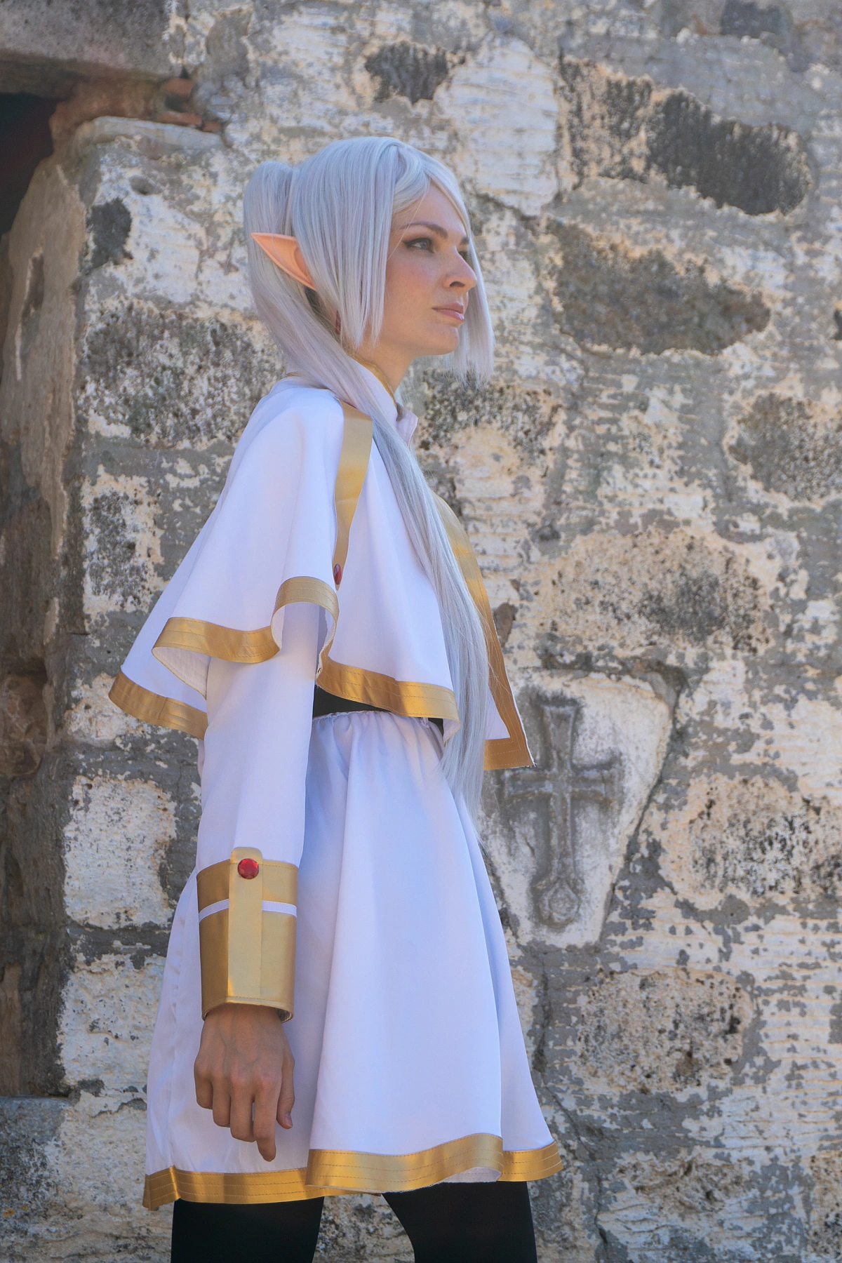 Frieren cosplay side profile against stone wall silver hair elf ears white gold mage dress gold trim detail soft daylight