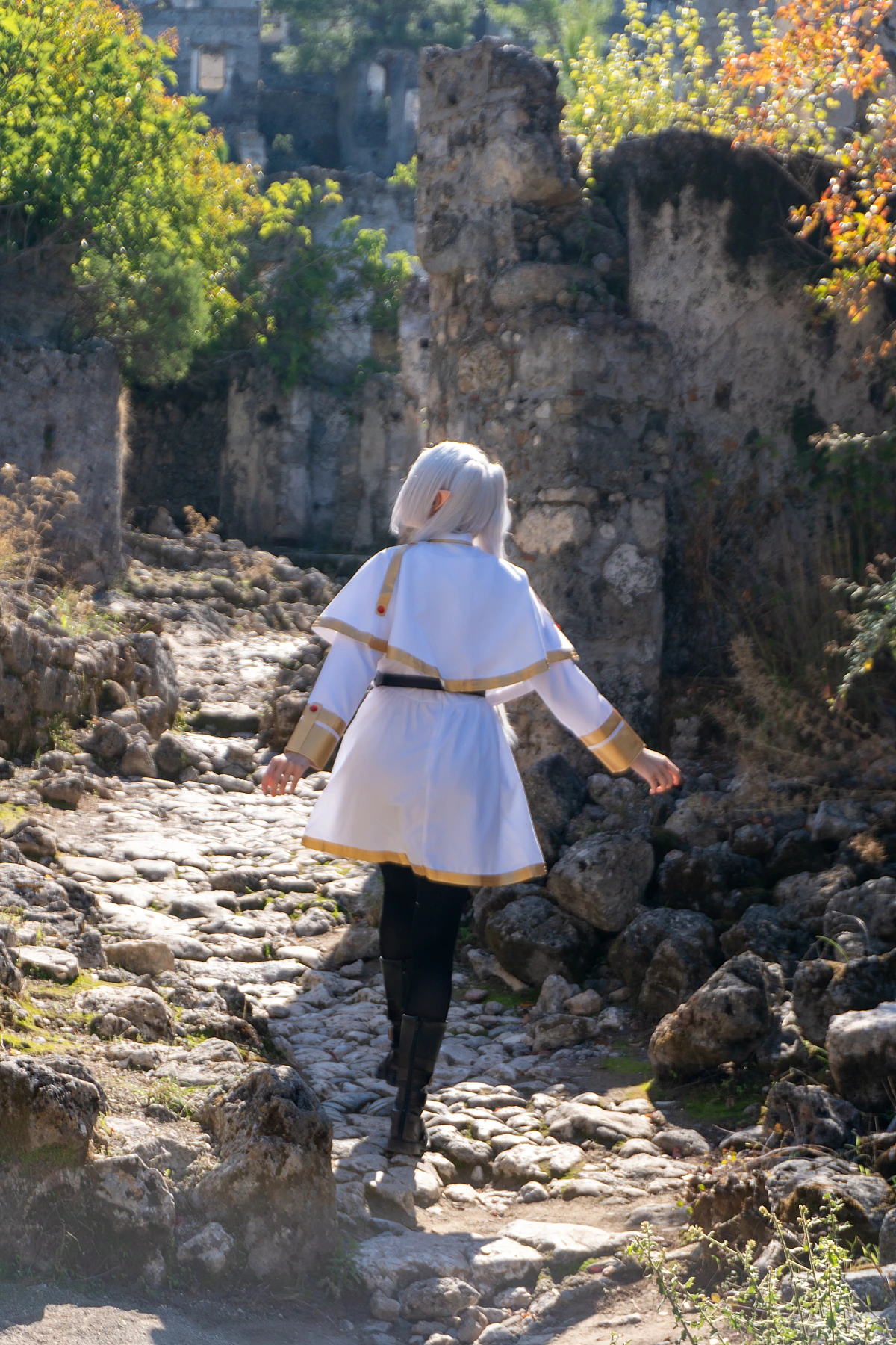 Frieren cosplay back view walking stone path silver hair elf ears white gold mage dress ancient ruins daylight