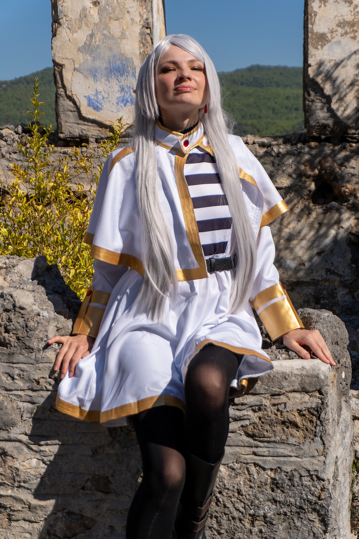 Frieren cosplay sitting stone ruins face lifted to sunlight silver hair elf ears white gold mage dress natural light