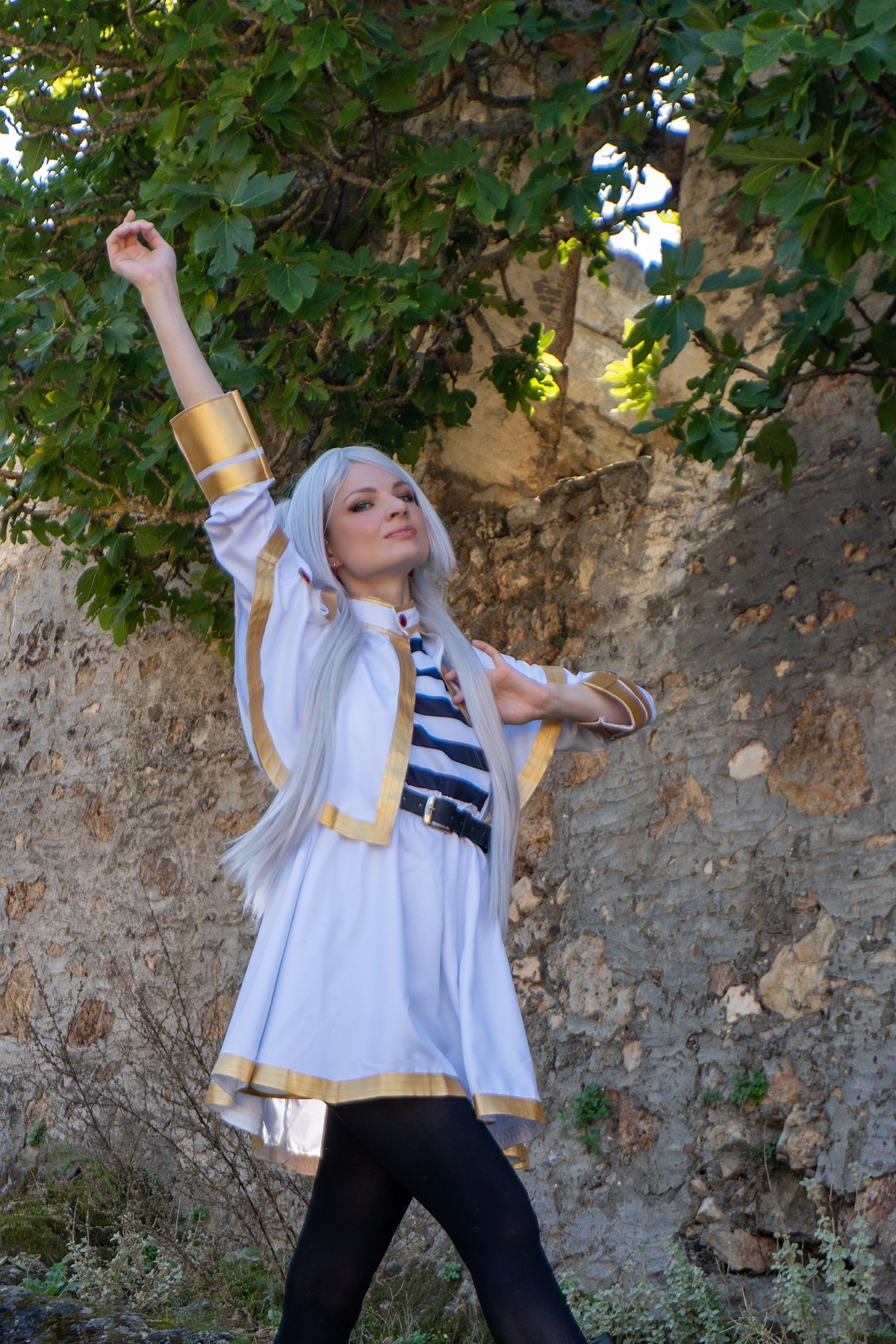 Frieren cosplay raised arm pose under green leaves silver hair elf ears white gold mage outfit stone wall ruins daylight