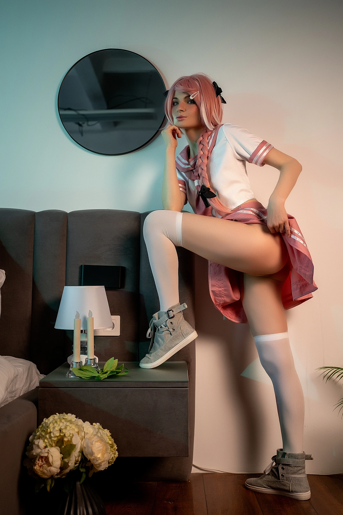 Astolfo trap cosplay with leg raised on nightstand in pink school uniform with white thigh-high socks and braided hair