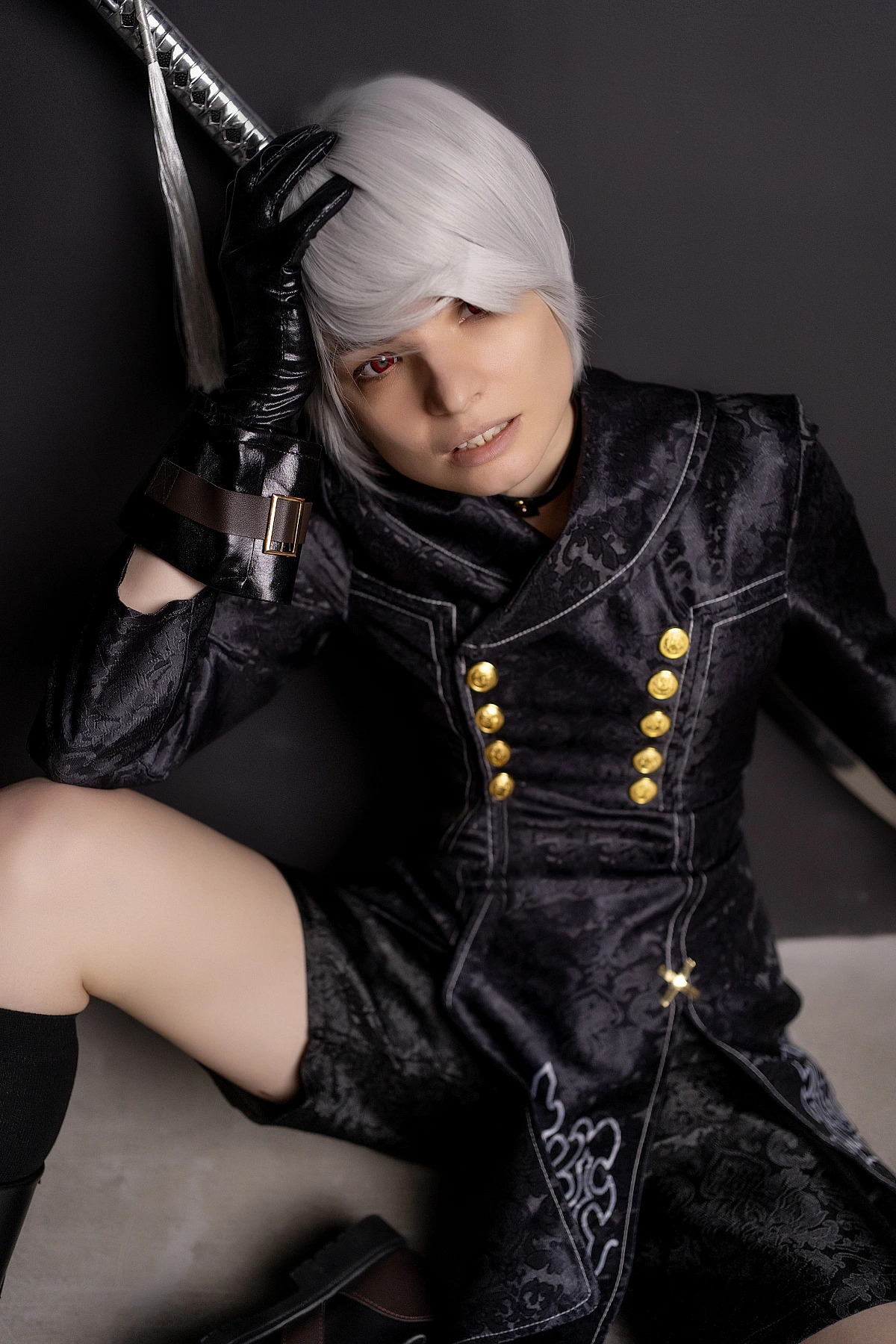 Close-up 9S cosplay portrait with silver hair, black YoRHa coat and katana hilt on dark background.