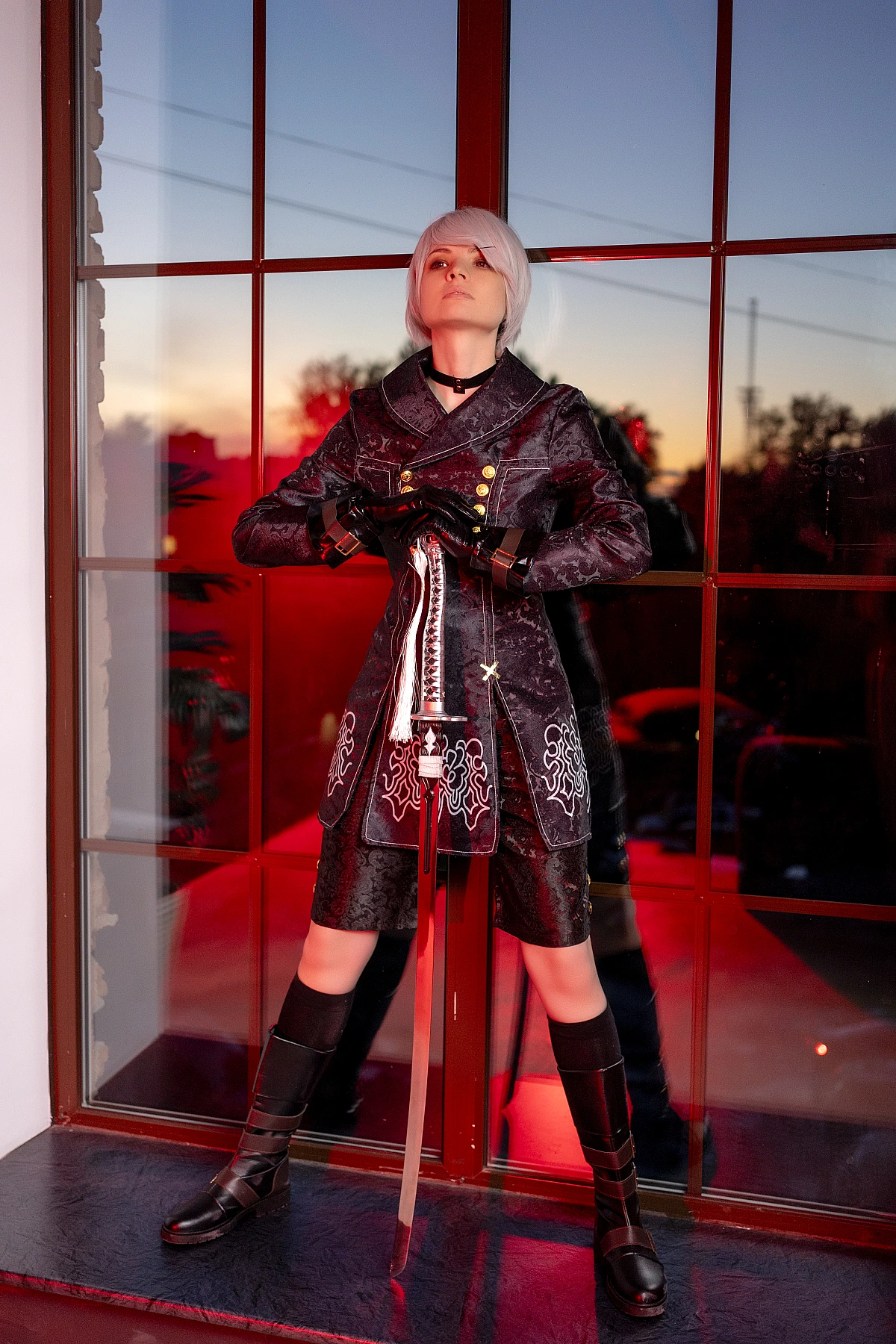 Full-body 9S cosplay standing by window at sunset with sword, black YoRHa coat and combat boots.