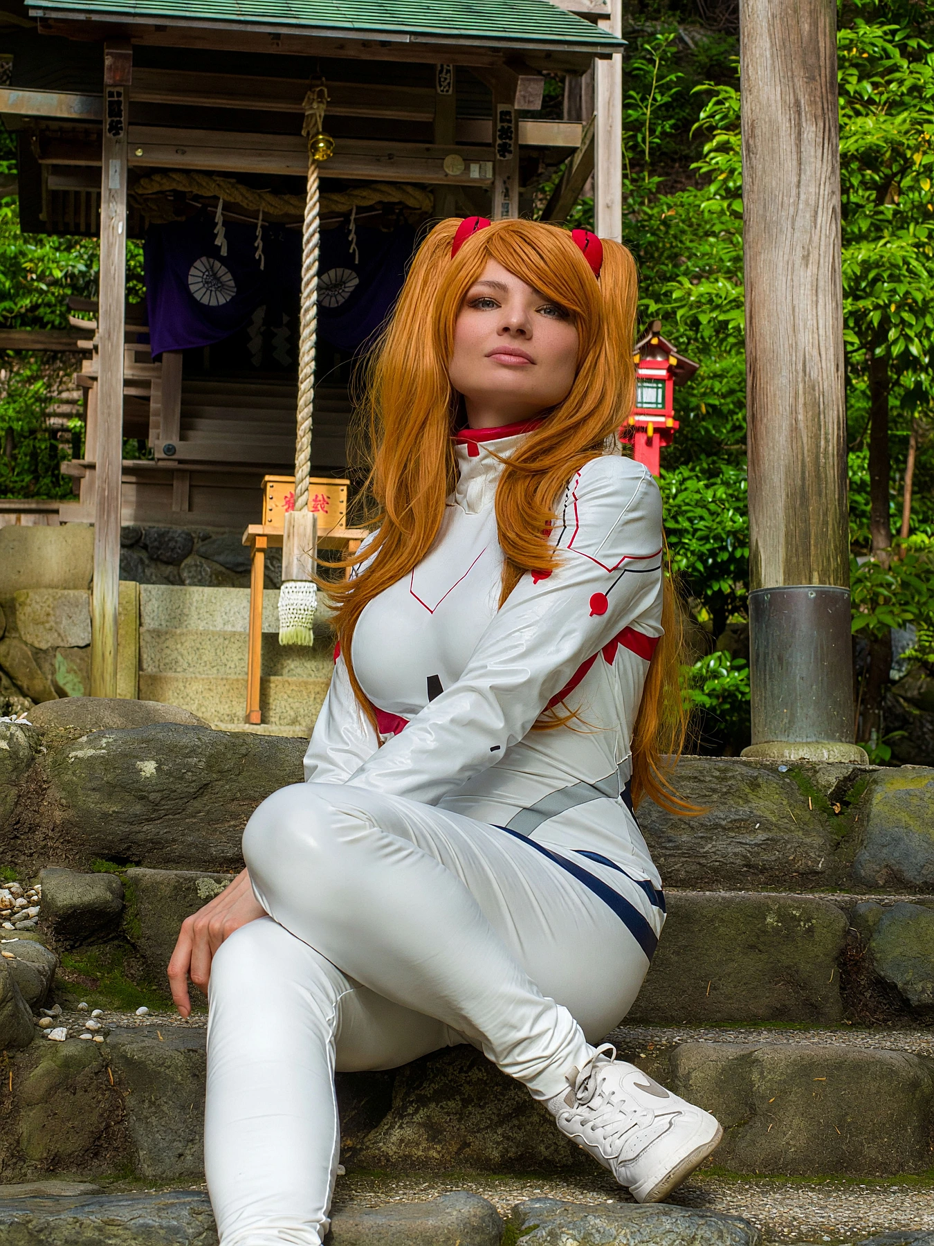 Asuka Langley cosplay seated near a Japanese shrine in a white Rebuild plugsuit with orange twin-tail wig