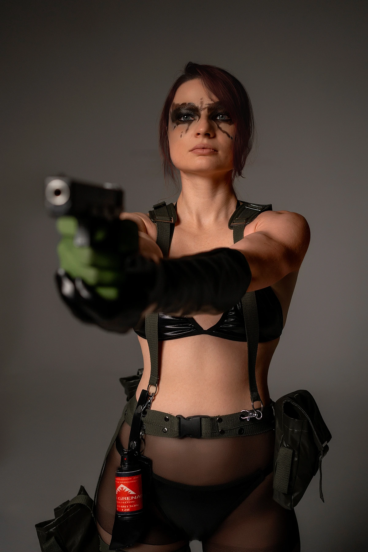 Quiet cosplay aiming a prop handgun in a dark studio with face paint, tactical gear and black outfit