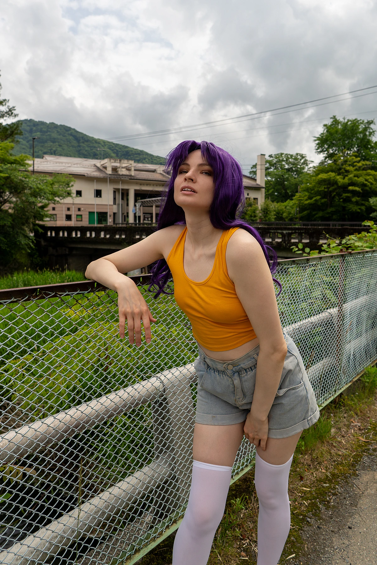 Misato Katsuragi casual cosplay posing near a river fence with purple hair, yellow top and mountain town background