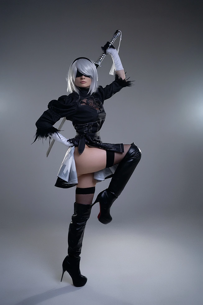 2B NieR Automata cosplay dynamic pose with raised leg and sword, silver hair, blindfold, black YoRHa dress and boots