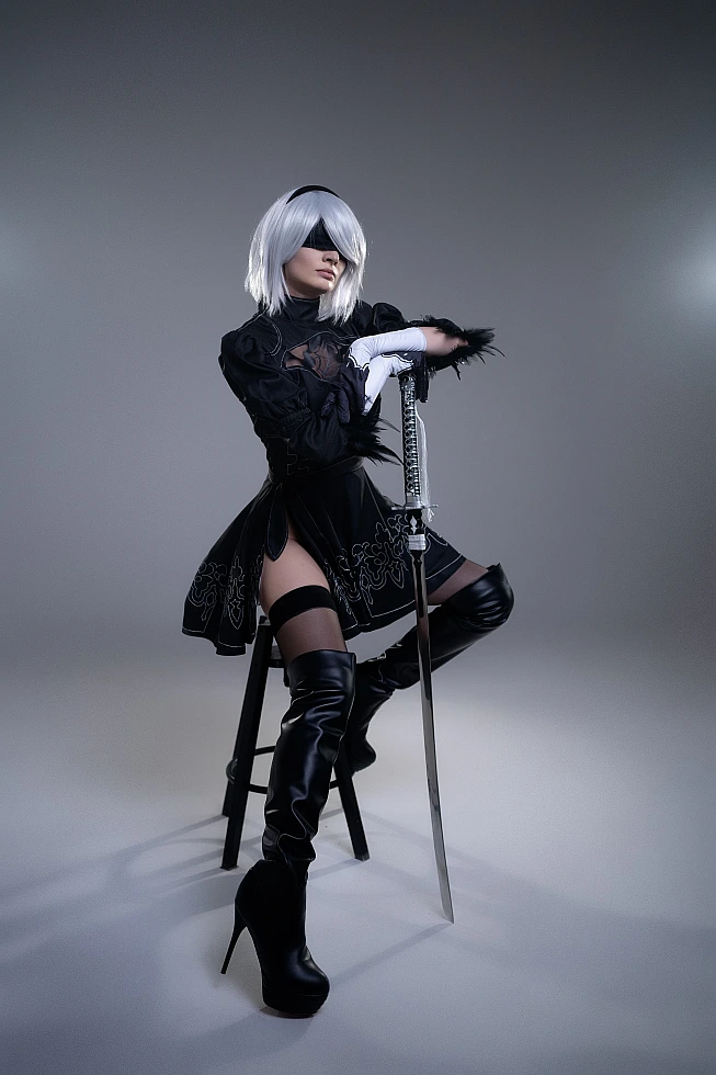 2B NieR Automata cosplay seated portrait with katana, silver hair, black blindfold, YoRHa black dress and thigh-high boots in studio