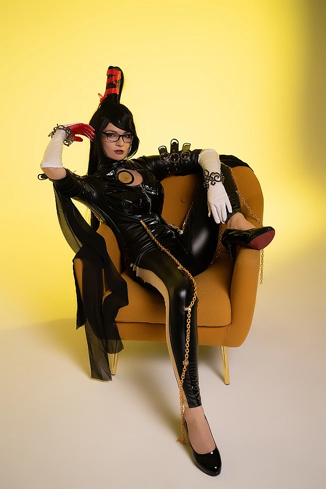 Bayonetta cosplay seated chair pose by Neka-Chi, glossy black bodysuit with gold chains, yellow studio background