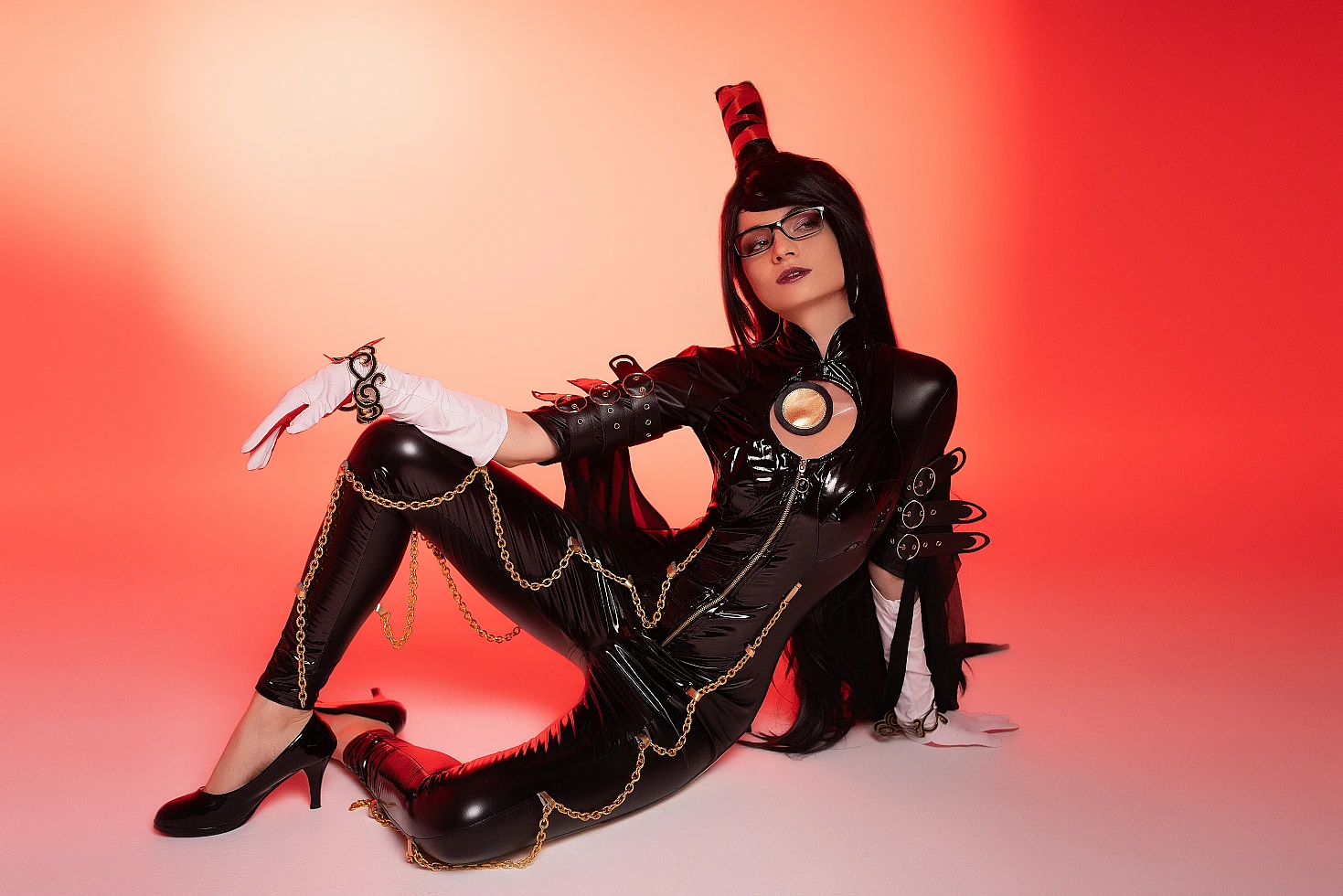 Bayonetta cosplay reclining floor pose by Neka-Chi, glossy black bodysuit with gold chain accents, red gradient studio lighting