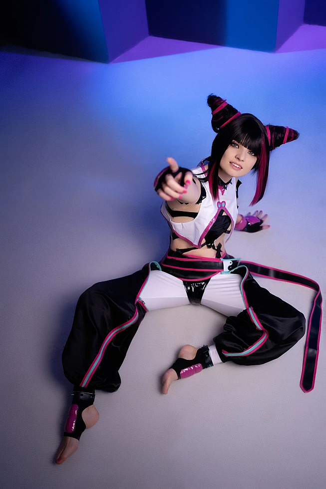 Juri Han cosplayer seated on studio floor reaching upward, barefoot, cool blue-violet neon background, horn buns, Street Fighter