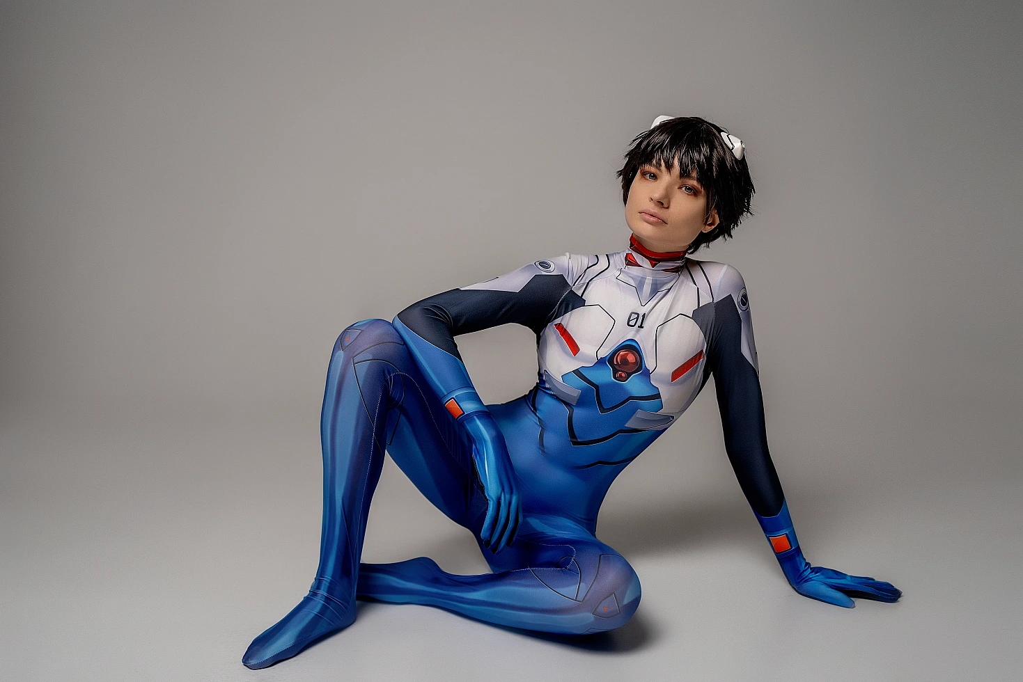 Shinji Ikari cosplay reclining floor pose, blue Evangelion plugsuit, anime crossplay photography, gray studio background