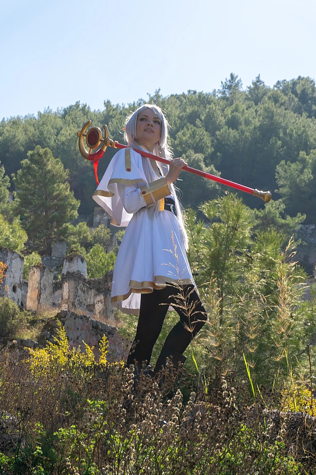 Frieren cosplay staff resting on shoulder standing tall silver hair elf ears white gold mage dress forest ruins daylight traveler