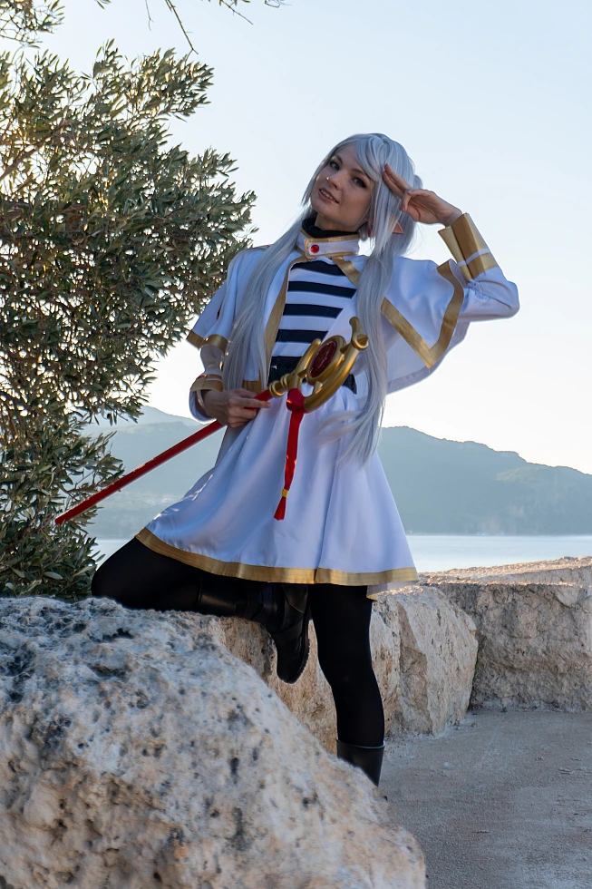 Frieren cosplay playful hand gesture near temple relaxed stance silver hair elf ears white gold mage dress staff seaside wall