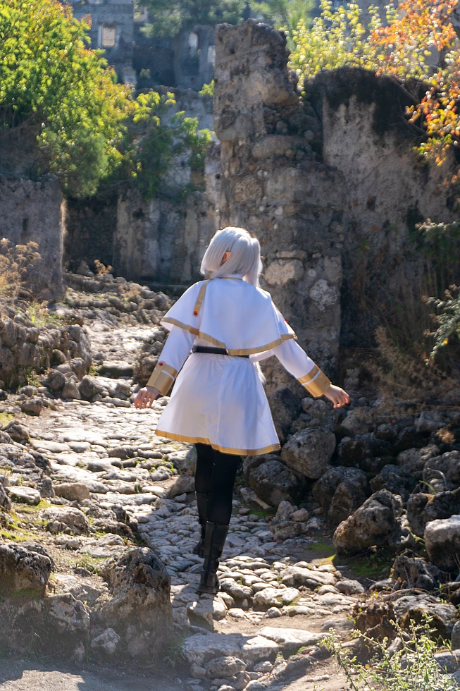 Frieren cosplay back view walking stone path silver hair elf ears white gold mage dress ancient ruins daylight