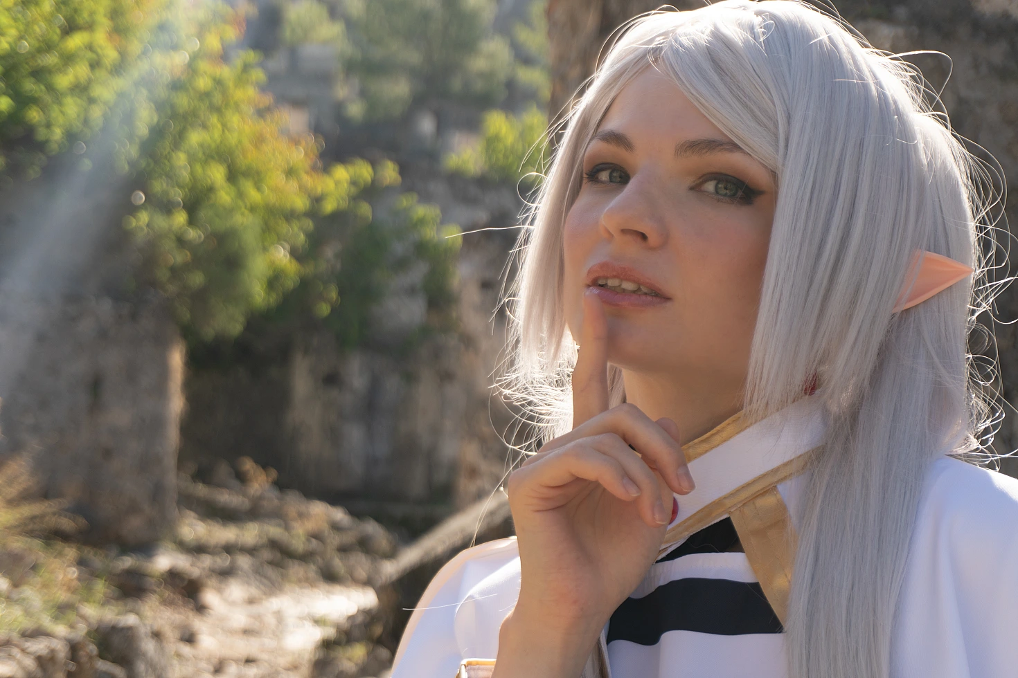 Frieren cosplay close portrait finger to lips gesture silver hair elf ears white gold costume warm sunlight ruins background