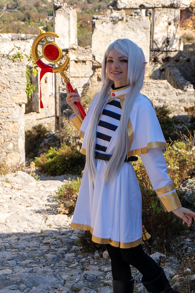 Frieren cosplay portrait holding staff gentle smile silver hair elf ears white gold mage outfit aged stone ruins daylight