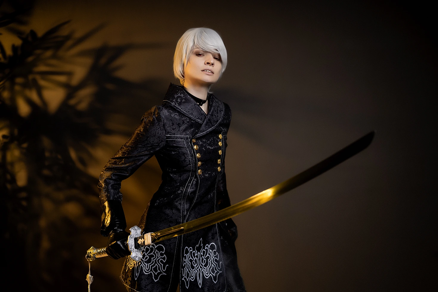 9S NieR:Automata cosplay holding katana in warm studio lighting, black YoRHa coat and white wig.