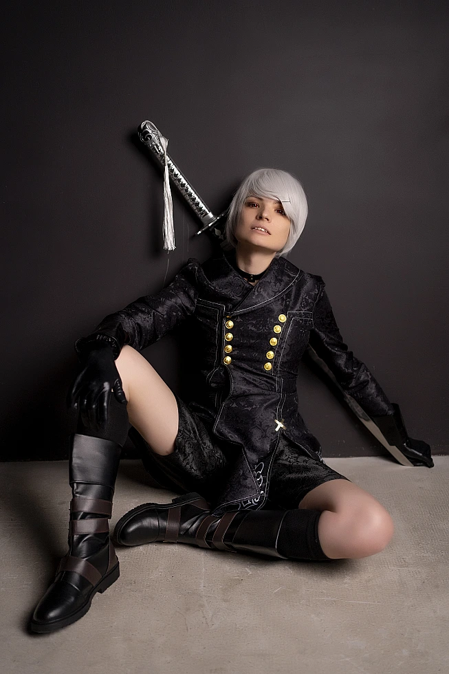 Seated 9S NieR:Automata cosplay in black YoRHa coat and white wig, leaning by dark wall with sword.