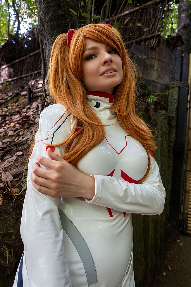 Close-up Asuka Langley cosplay in a glossy white Rebuild plugsuit with orange twin-tail wig beside trees and bamboo