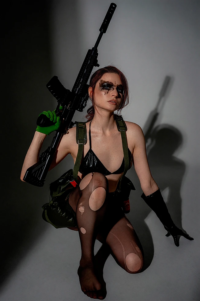 Quiet cosplay kneeling in a dark studio with a prop rifle, tactical gear and face paint
