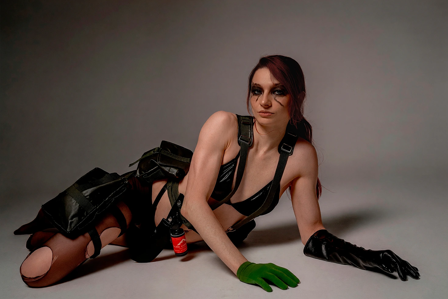 Quiet cosplay reclining on the studio floor with tactical gear, green glove and dark character makeup