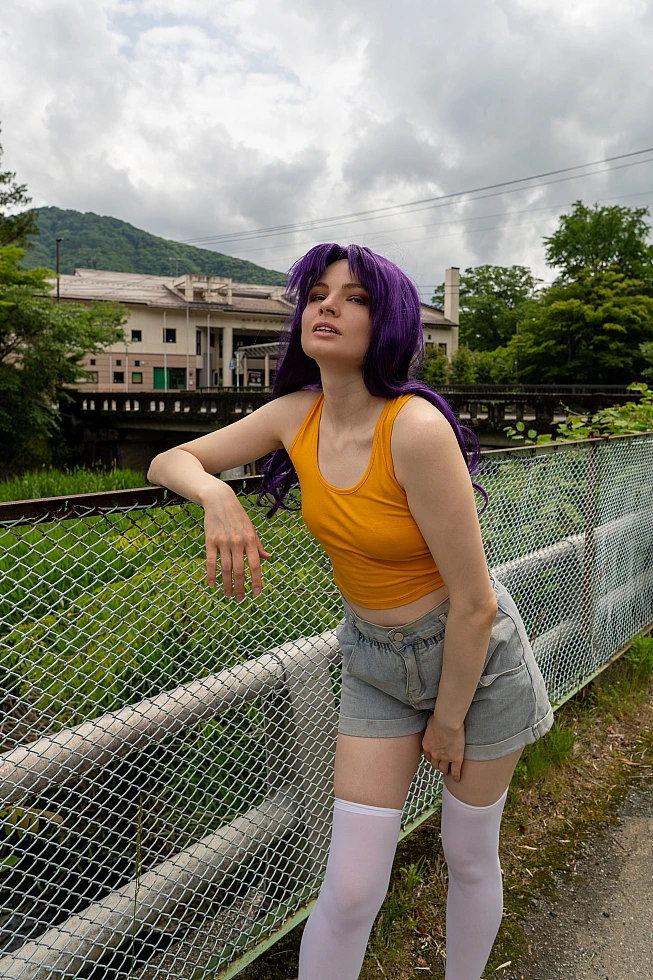 Misato Katsuragi casual cosplay posing near a river fence with purple hair, yellow top and mountain town background