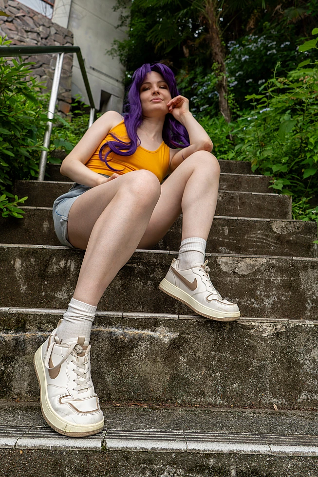 Misato Katsuragi casual cosplay seated on outdoor stone steps with purple hair, yellow top, denim shorts and sneakers