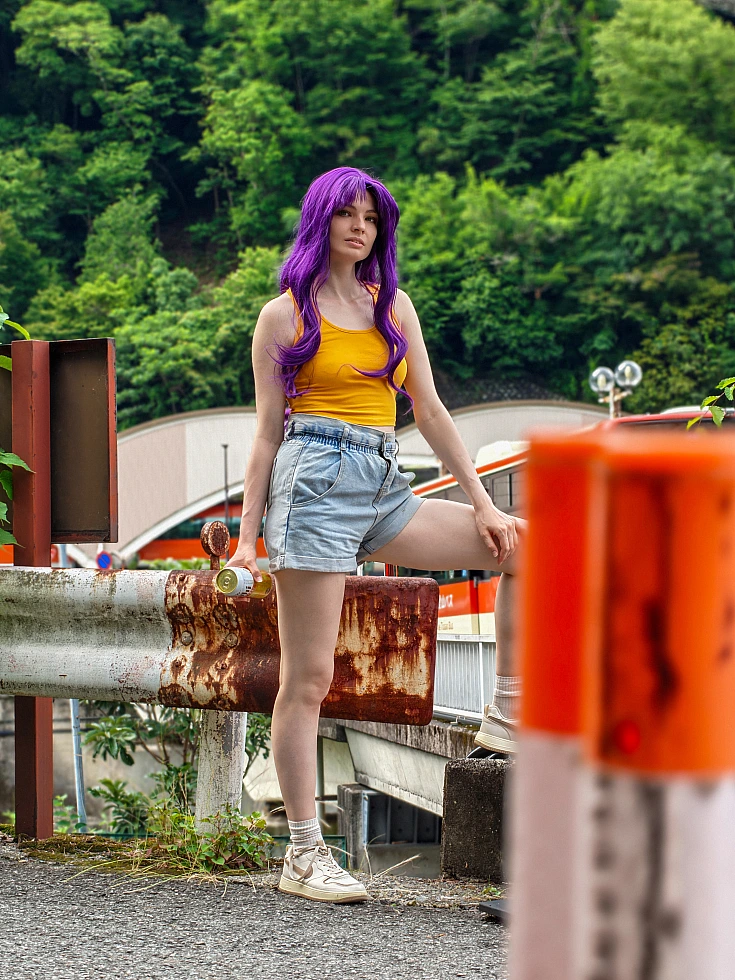 Misato Katsuragi casual cosplay standing by a mountain road guardrail with purple hair, yellow top and denim shorts