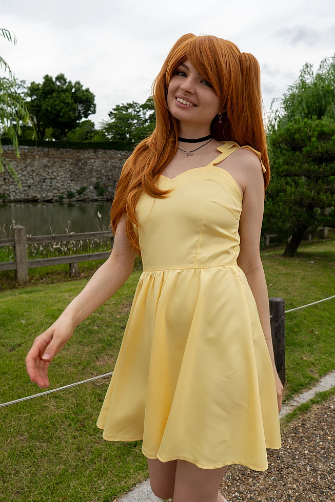 Asuka Langley casual cosplay smiling beside a Japanese castle moat in a yellow summer dress with long orange twin-tail hair