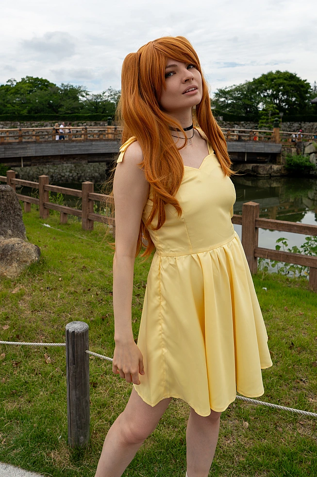 Asuka Langley casual cosplay posing beside a Japanese castle moat in a yellow dress with long orange twin-tail hair