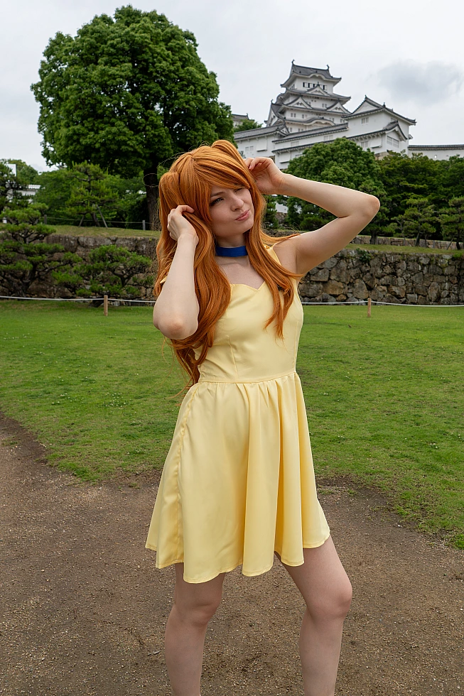 Asuka Langley casual cosplay posing in a yellow summer dress with Himeji Castle and green gardens behind her