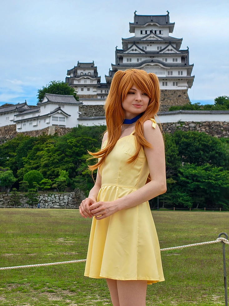 Asuka Langley cosplay in a yellow summer dress standing in front of Himeji Castle with long orange twin-tail hair