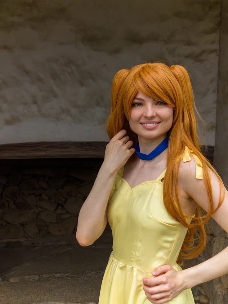 Asuka Langley cosplay in a yellow summer dress posing beside a traditional stone wall with orange twin-tail hair
